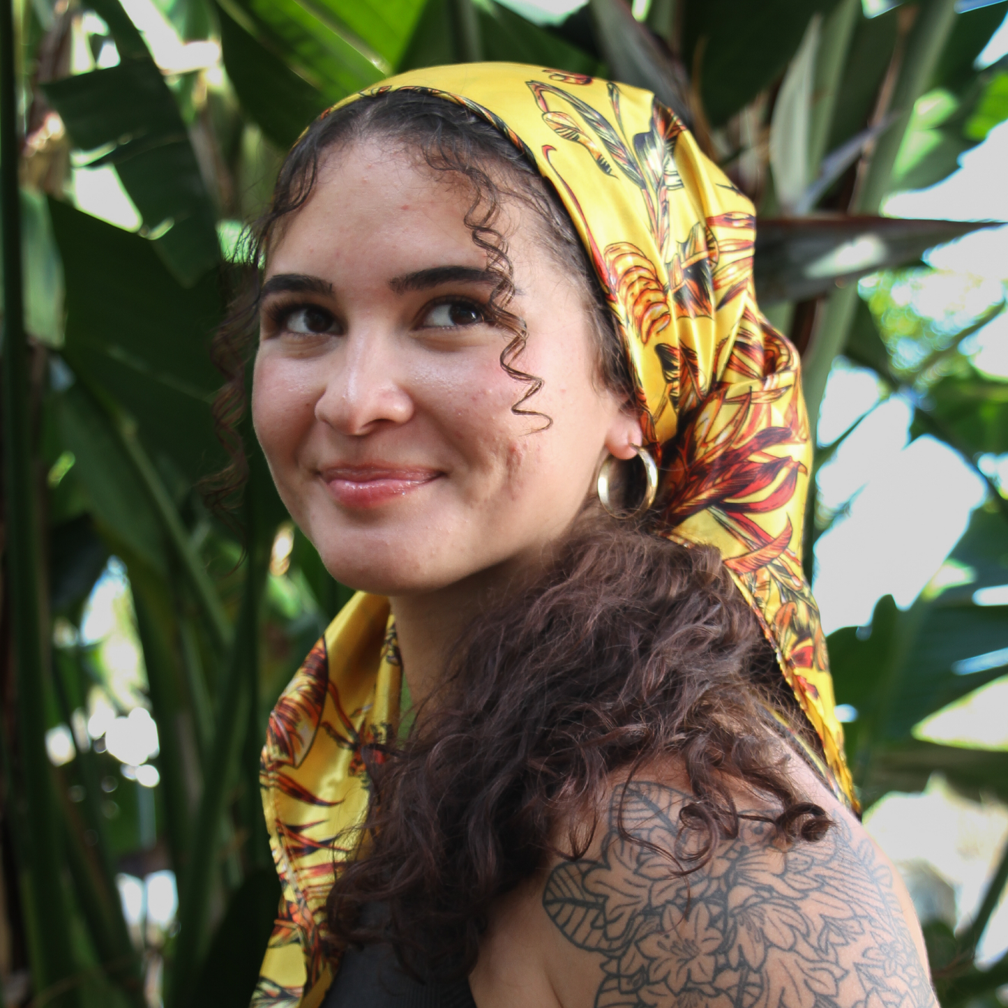 Woman wearing a silky yellow Broadus Collection head scarf with tropical print, smiling with curly hair and floral tattoo visible