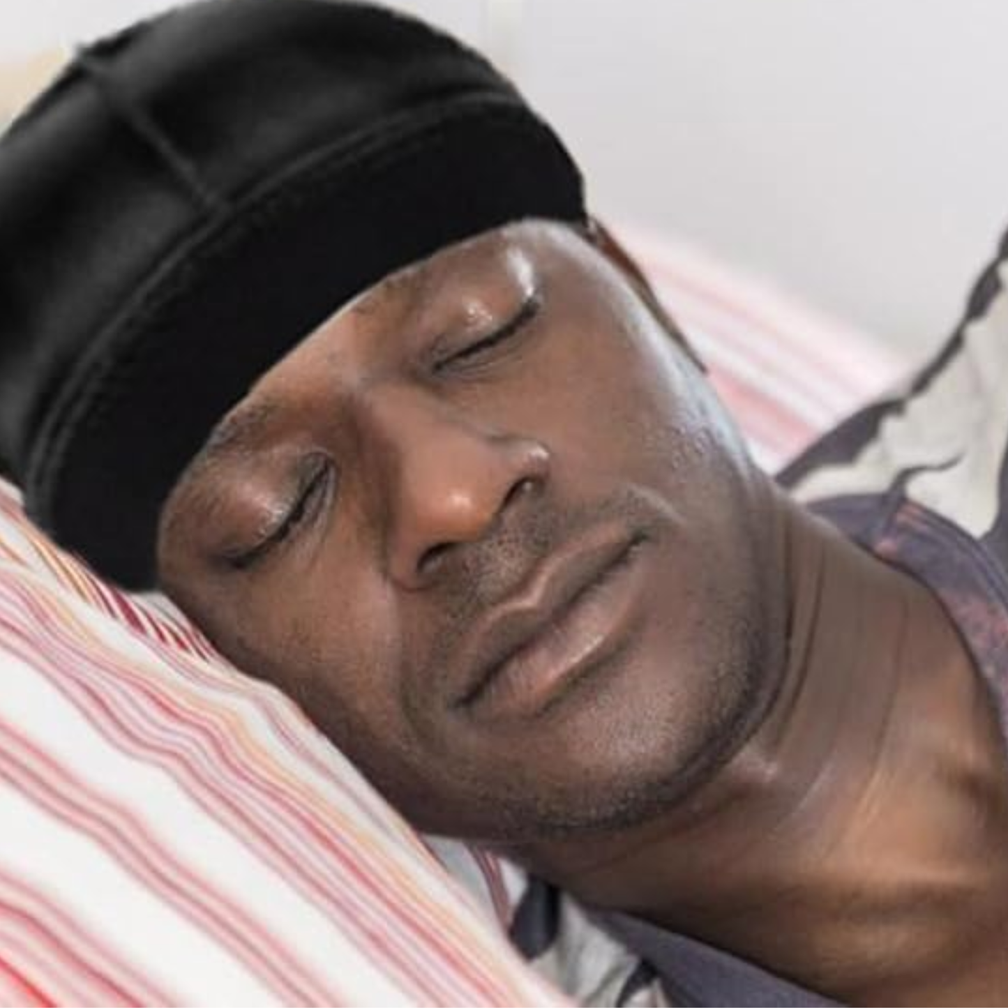 Man wearing black Mr. Durag Stocking Wave Cap with wide band, resting on striped pillow with eyes closed.
