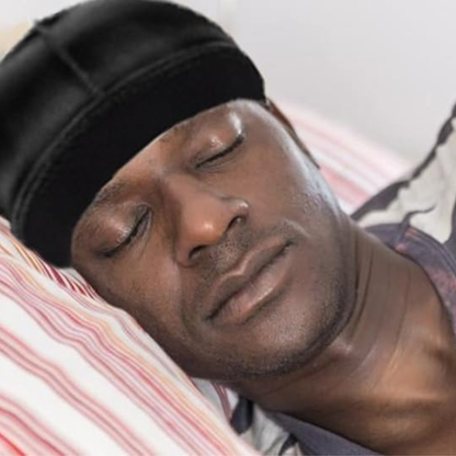 Man wearing black Mr. Durag Stocking Wave Cap with wide band, resting on striped pillow with eyes closed.