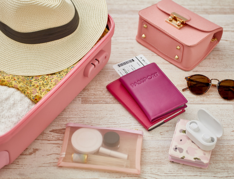 Open pink suitcase with straw hat, floral clothing, pink purse, sunglasses, passports, and travel-sized toiletries on wooden surface