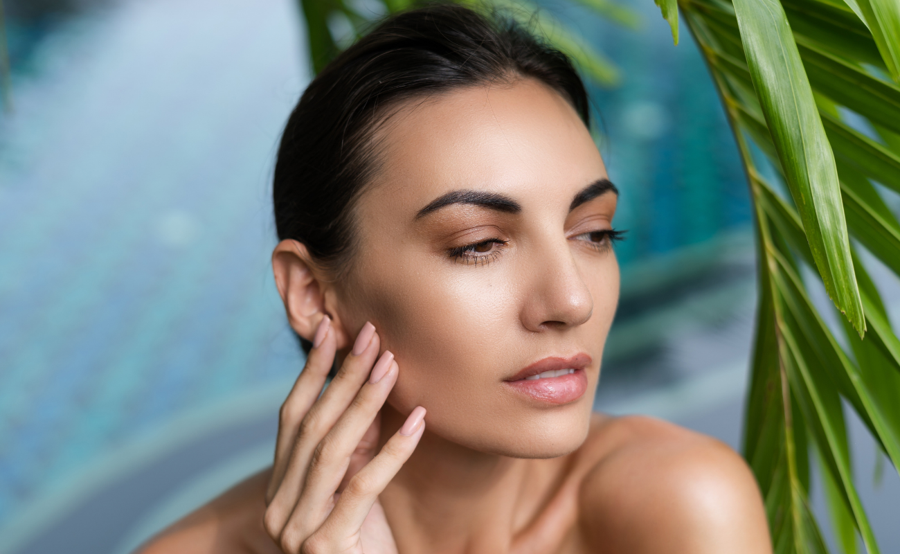 Close-up of a woman with smooth skin gently touching her face surrounded by green palm leaves with blurred blue background