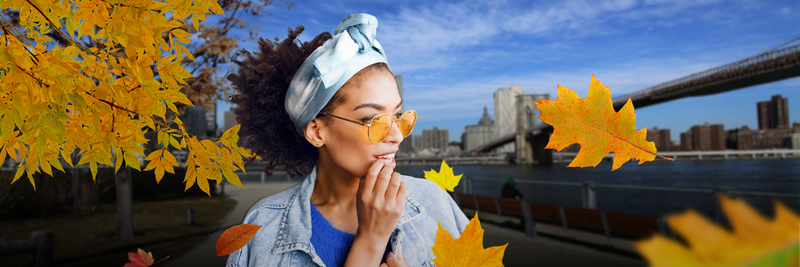 Young woman wearing sunglasses and headscarf standing by river with autumn leaves and city skyline in background