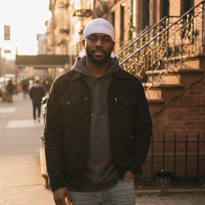 A man standing outdoors in soft evening light wearing a Mr. Durag Solid Durag, 1 Count, White styled under a dark jacket and hoodie, demonstrating a sleek look for 360 waves.