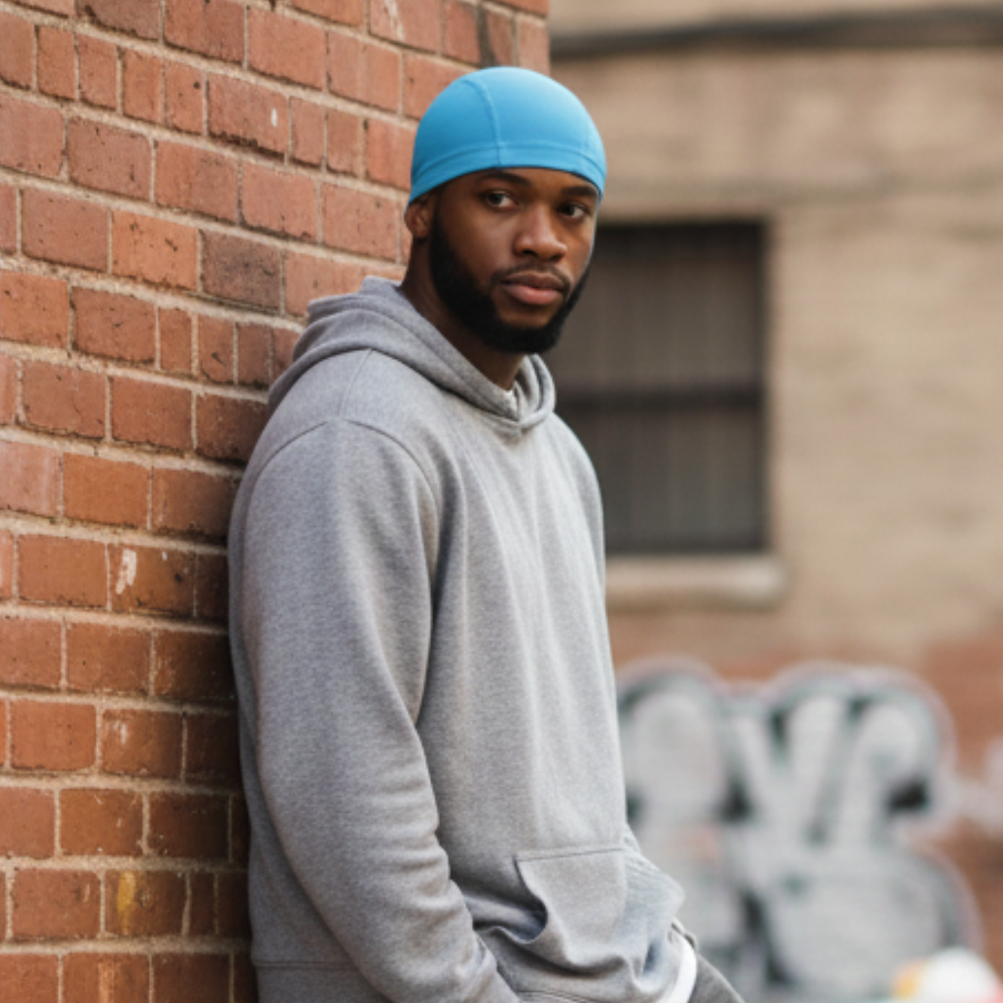 A man wearing Mr. Durag Spandex Dome Cap, 1 Count, Assorted Colors in bright blue, standing against a brick wall outdoors, featuring smooth, breathable spandex for comfortable wave training.