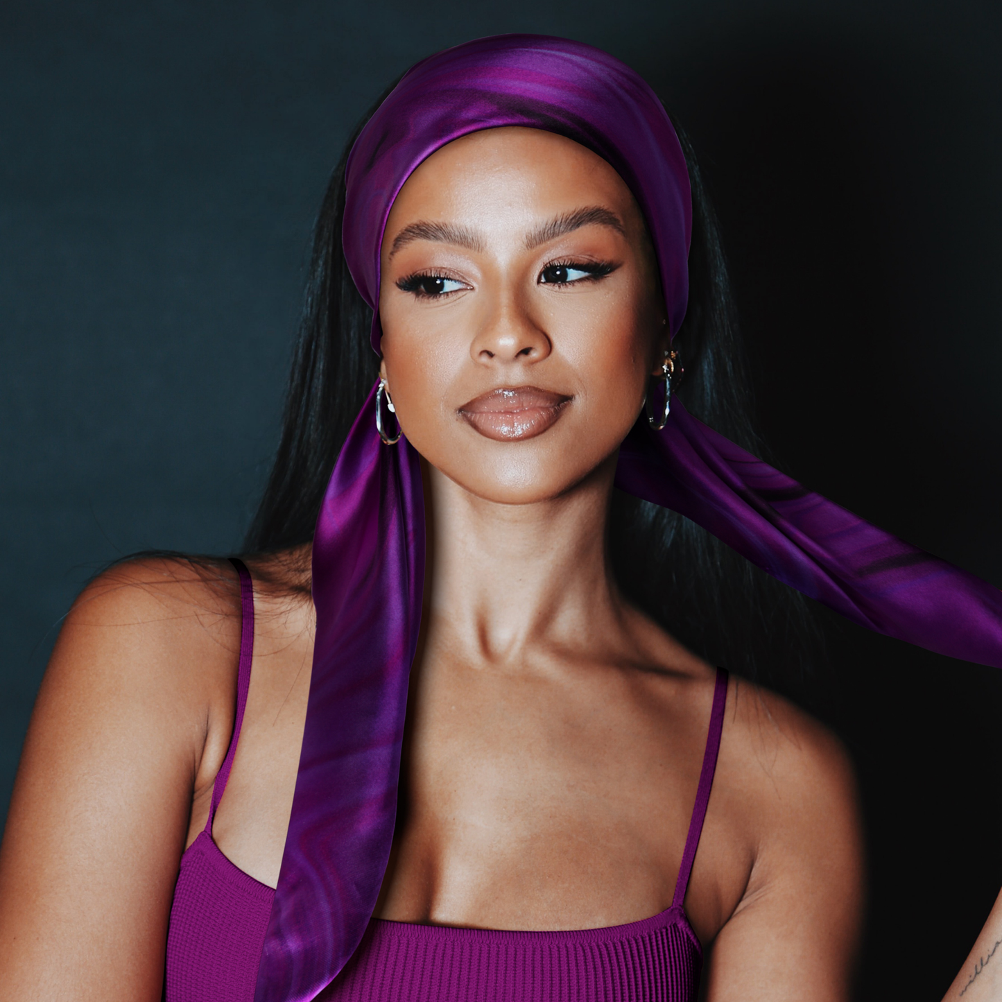 A confident woman with glossy makeup wears the Broadus Collection Headwrap Scarf by Shante & Snoop Dogg, Purple Butterfly, styled elegantly over long dark hair against a dark background.