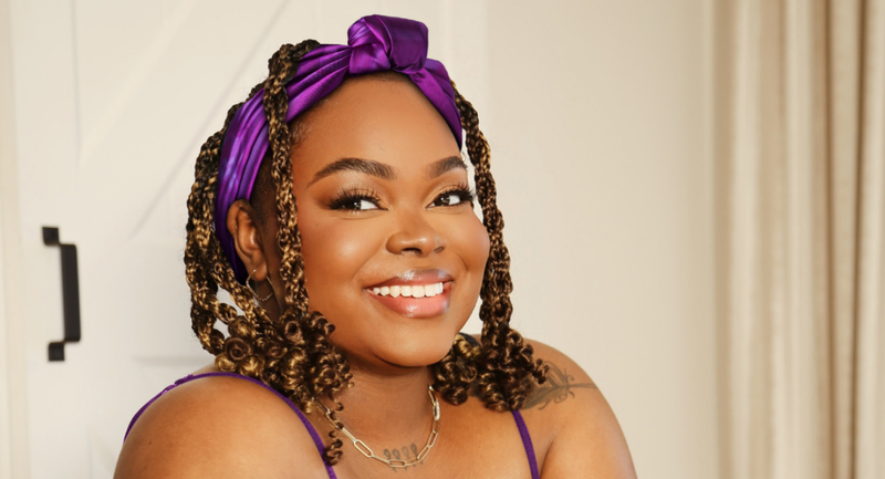 Smiling woman with curly hair wearing a purple headband and tank top sitting indoors against neutral background