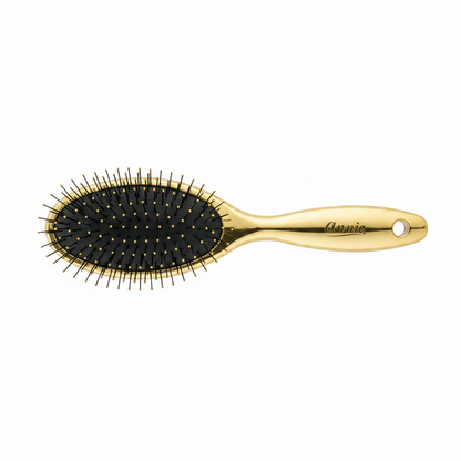 Gold-colored oval hairbrush with flexible bristles and cushioned black base, featuring an ergonomic handle engraved with the brand name for gentle detangling of textured hair.