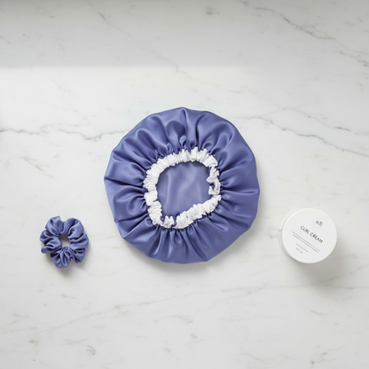 Flat-lay of a medium-sized purple satin bonnet with white elastic trim, a matching purple satin scrunchie, and a white jar of curl cream on a marble surface, styled for textured hair care.